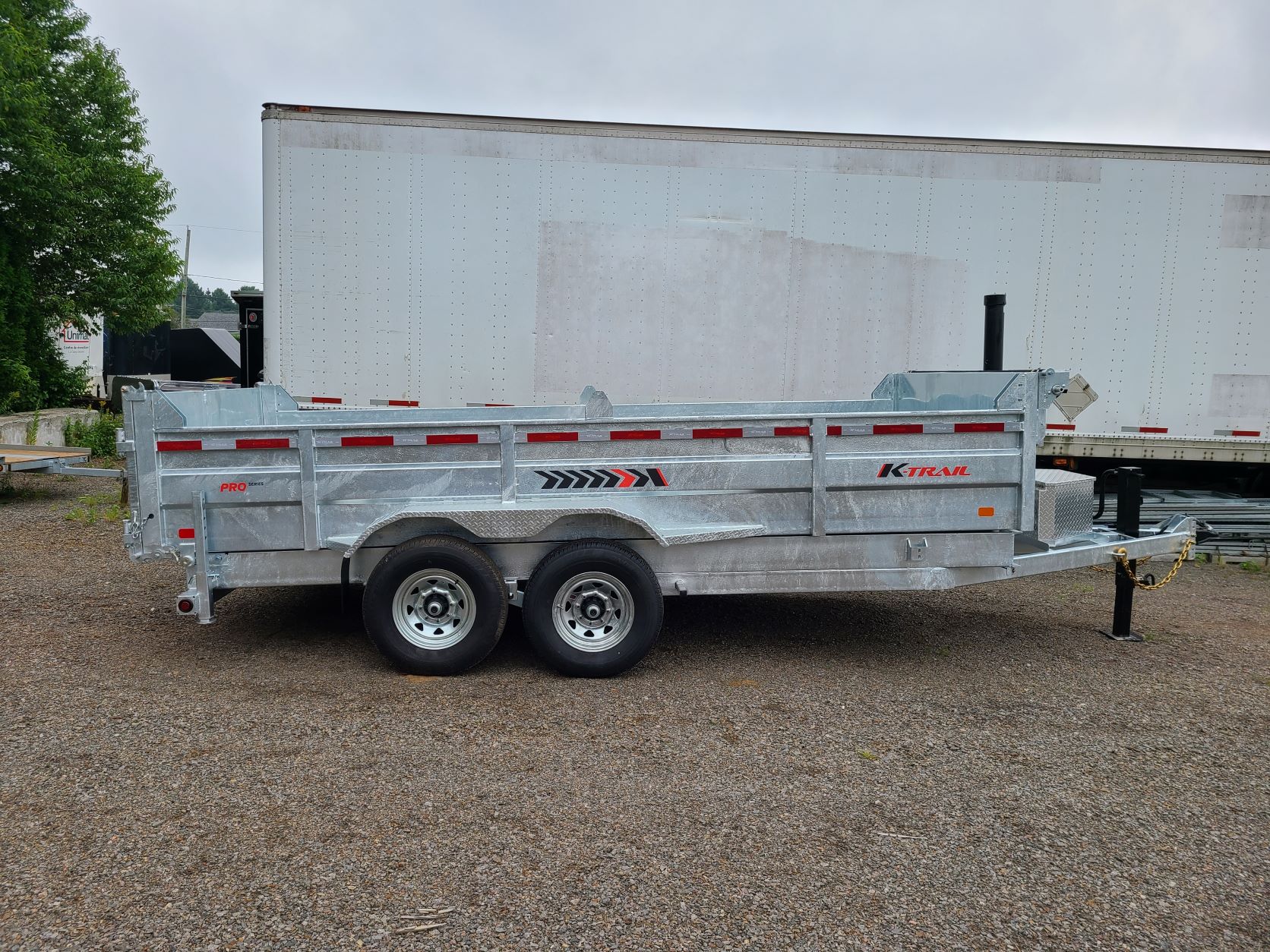 Une remorque à benne basculante est garée dans un terrain en gravier devant une remorque blanche.