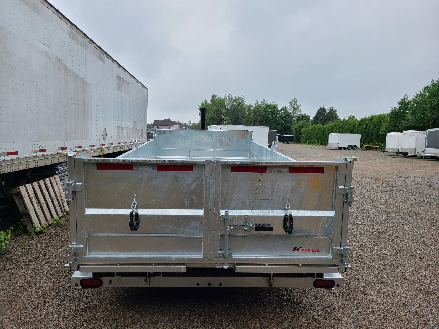 Une remorque à benne basculante est garée sur un terrain en gravier à côté d'un bâtiment.