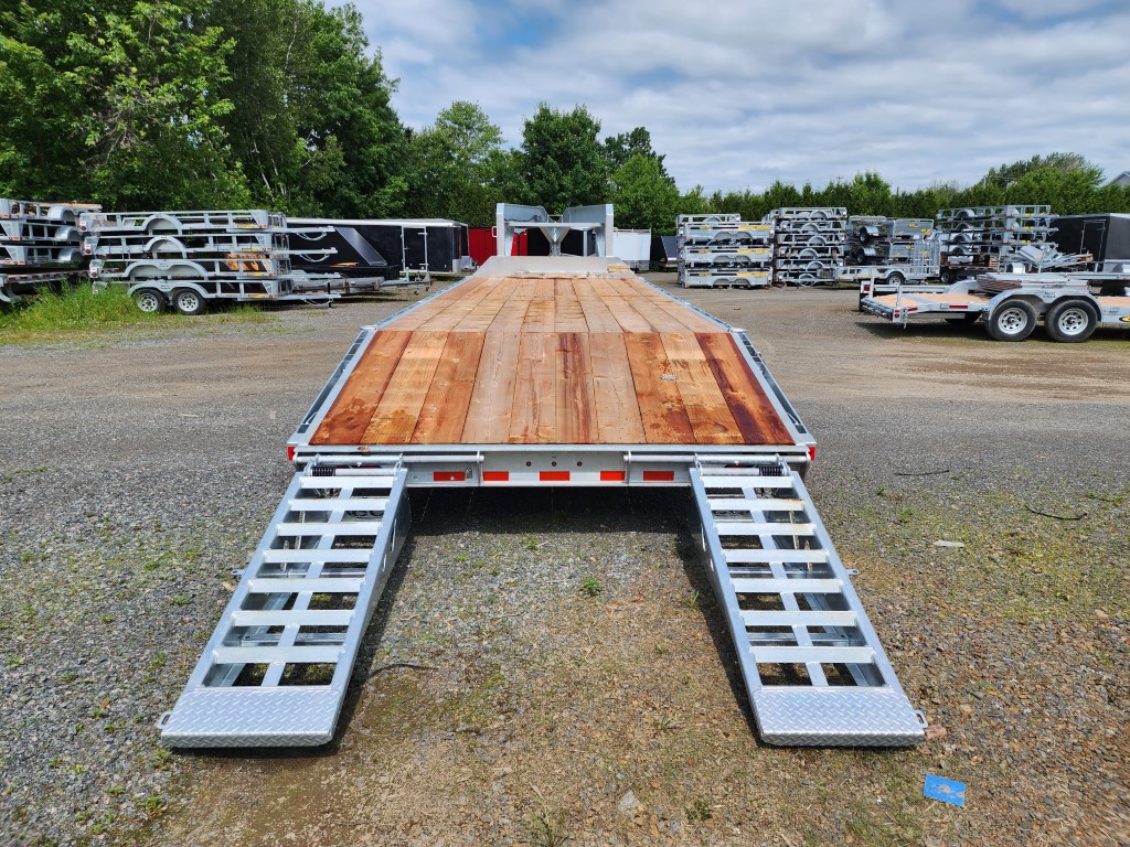 Une remorque à plateau avec rampes est garée sur un terrain en gravier.