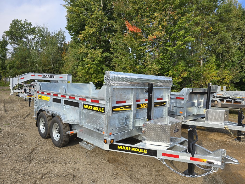 Une remorque à benne basculante est garée sur un terrain en terre avec des arbres en arrière-plan.
