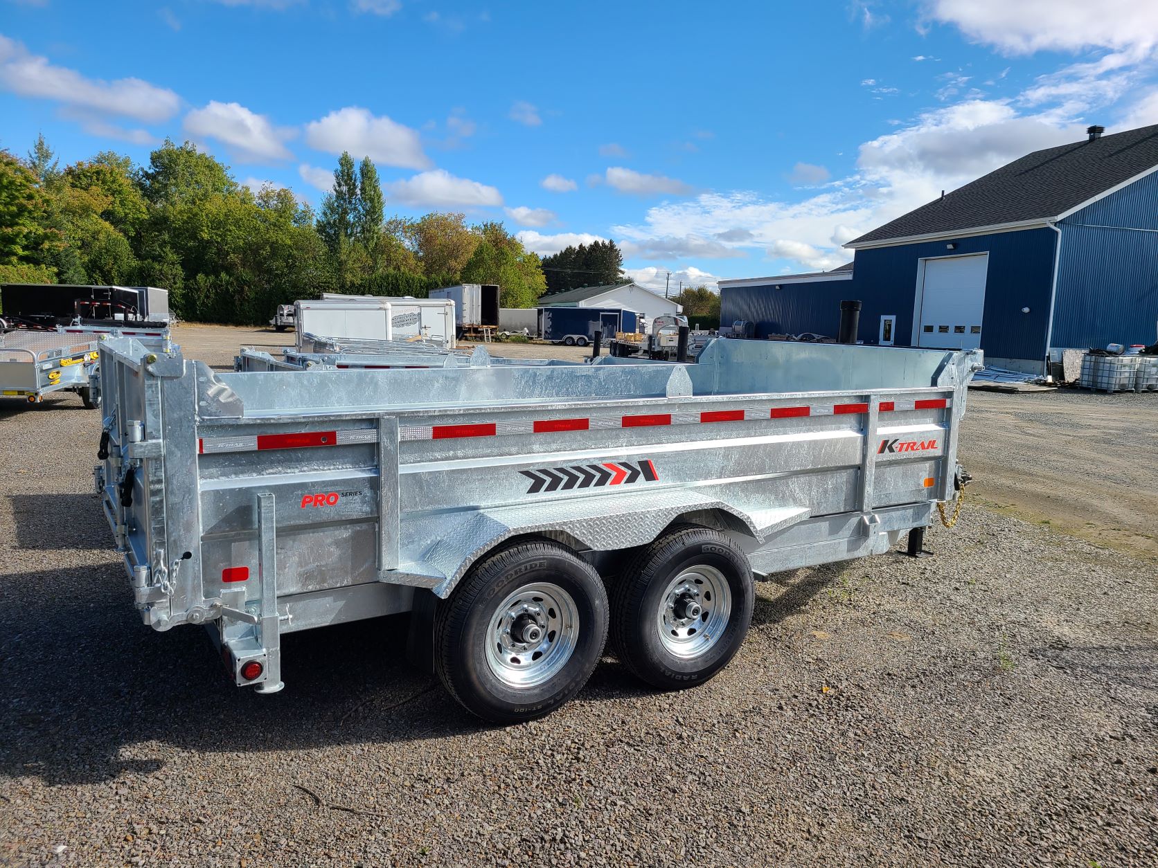 Une remorque à benne basculante est garée sur un terrain en gravier devant un bâtiment.